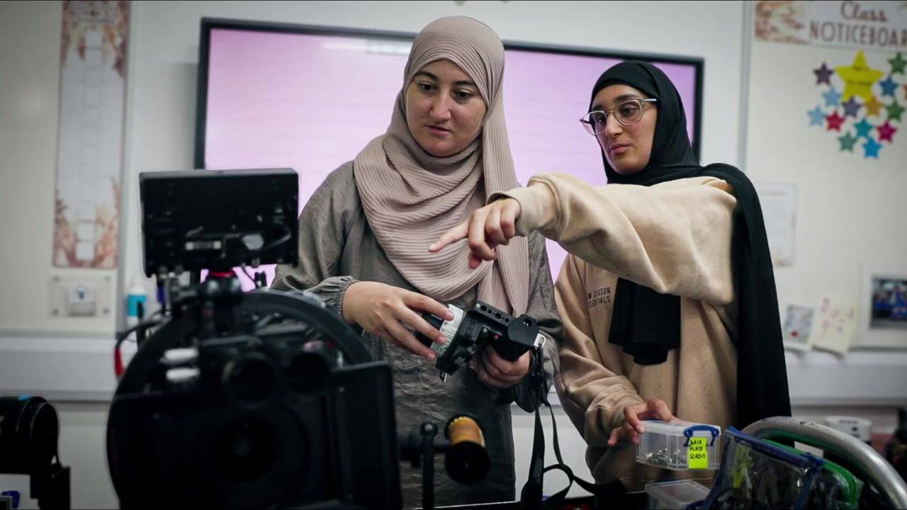 Two women in hijabs work on a built camera together, one pointing out a control on the body while the other holds a handset.