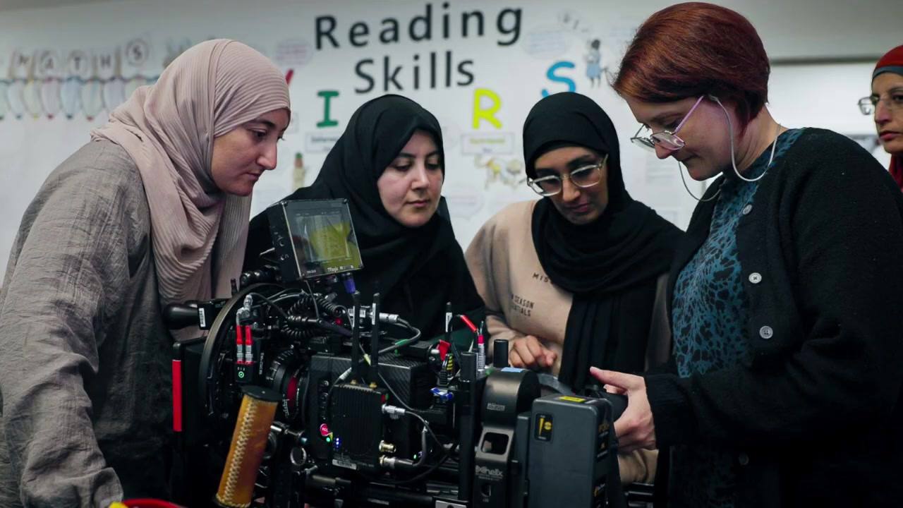 Four women, three of them in hijabs, lean in around a built camera and its monitor in a school classroom, a teacher among them, with a reading-skills display on the wall behind.