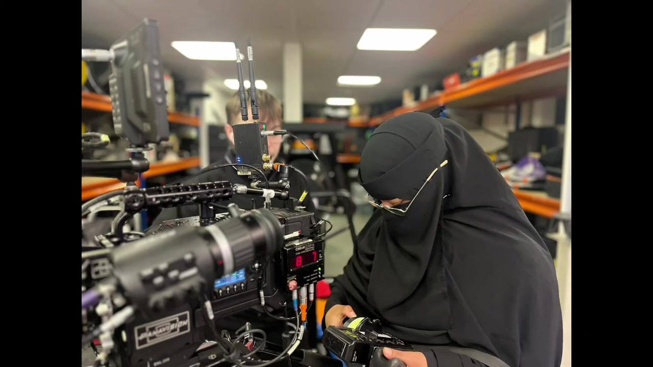 A woman in a niqab bends to look closely along the side of a large digital cinema camera on a bench in a kit store, racking shelves behind her.