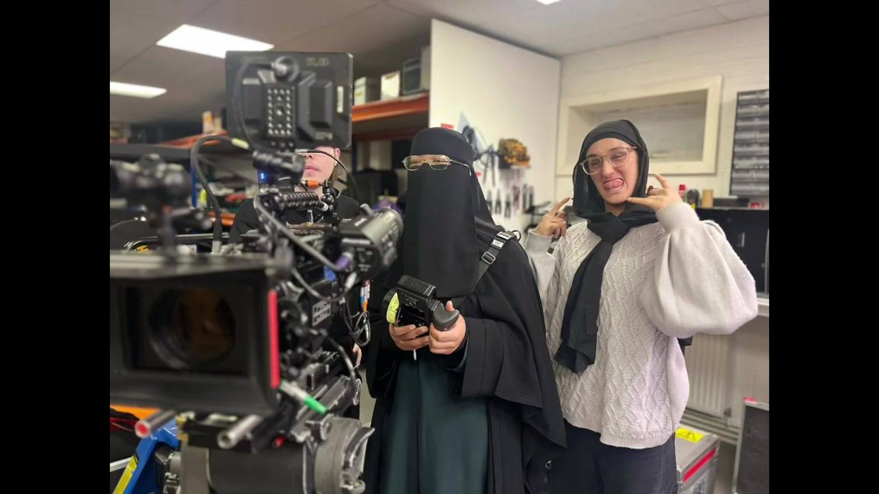 Two women stand in a kit store beside a fully built camera, one in a niqab holding a handset, the other laughing beside her.