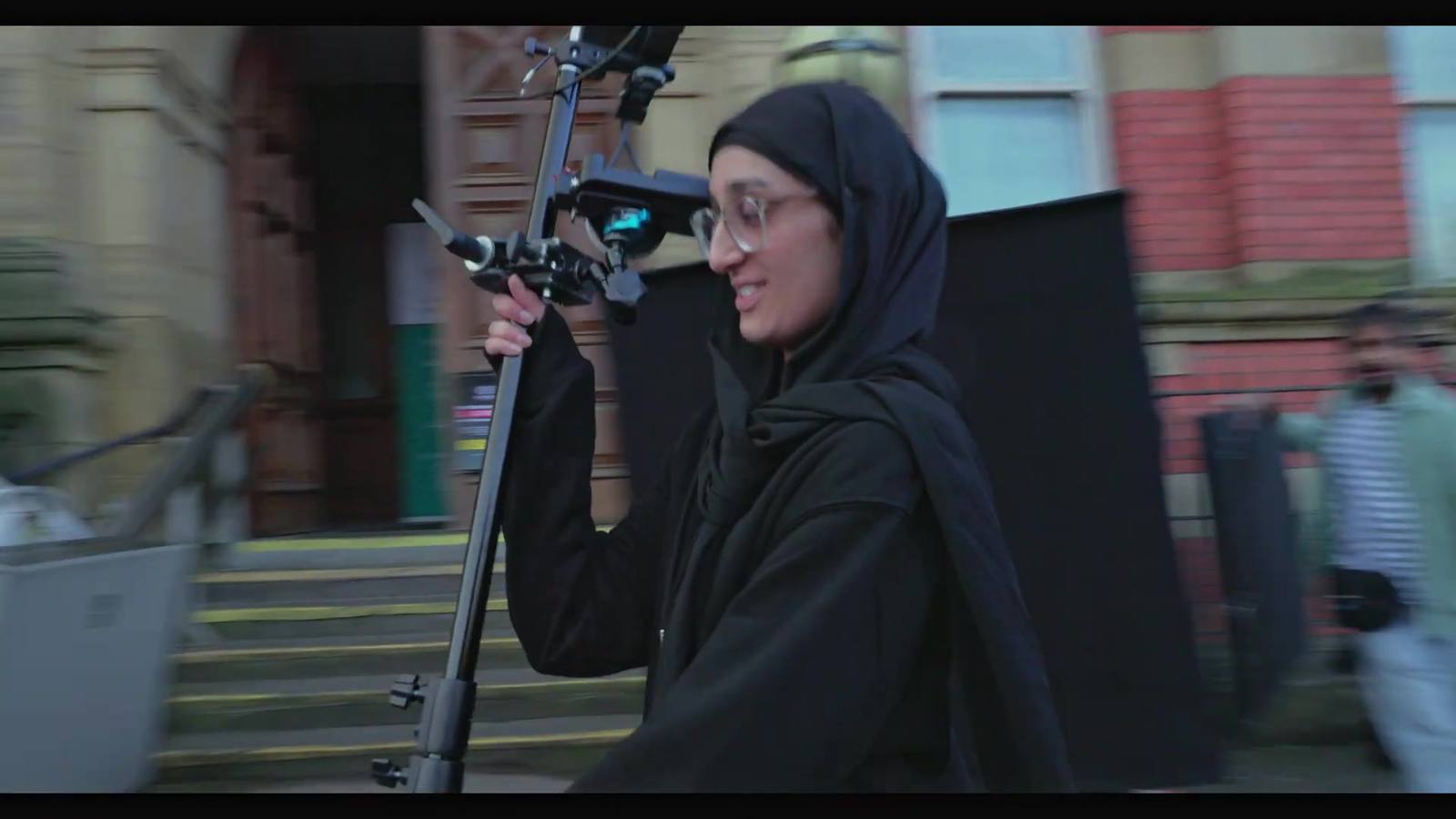 A woman in a hijab and glasses lifts a boom pole into position on a location street, smiling, with the unit and a set of steps behind her.