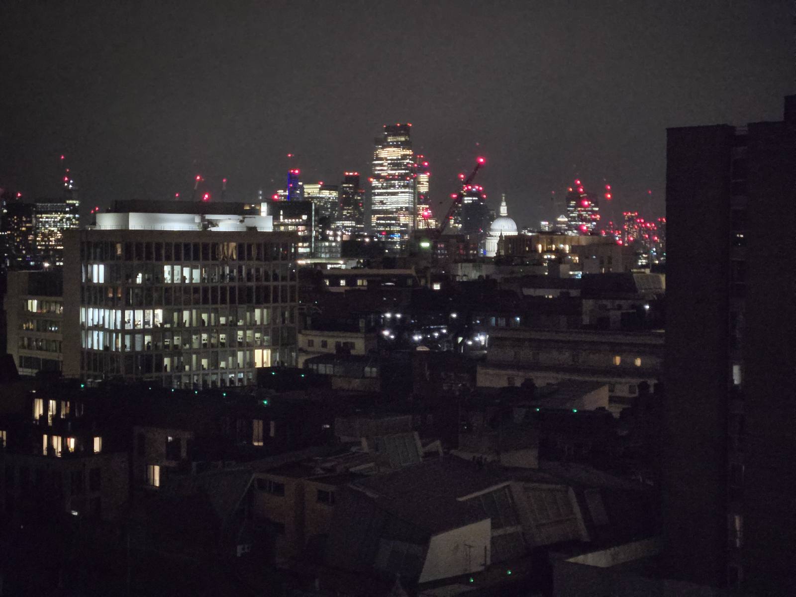 The London skyline at night seen across low rooftops, the towers of the City lit and a residential block in the foreground.