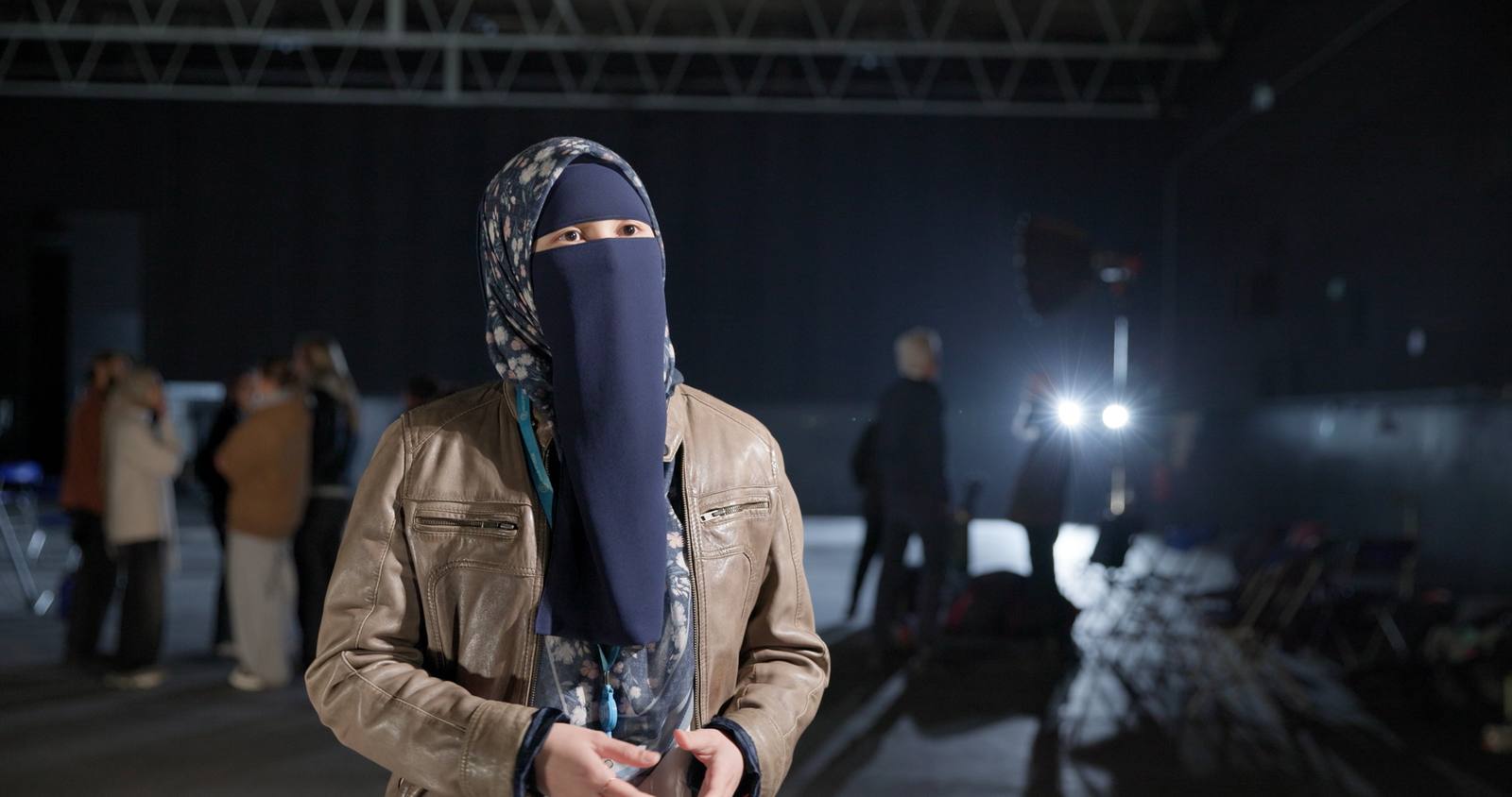 A participant in a niqab and a leather jacket stands on the stage floor at 3 Mills, crew working behind her and a lamp burning off to the right.