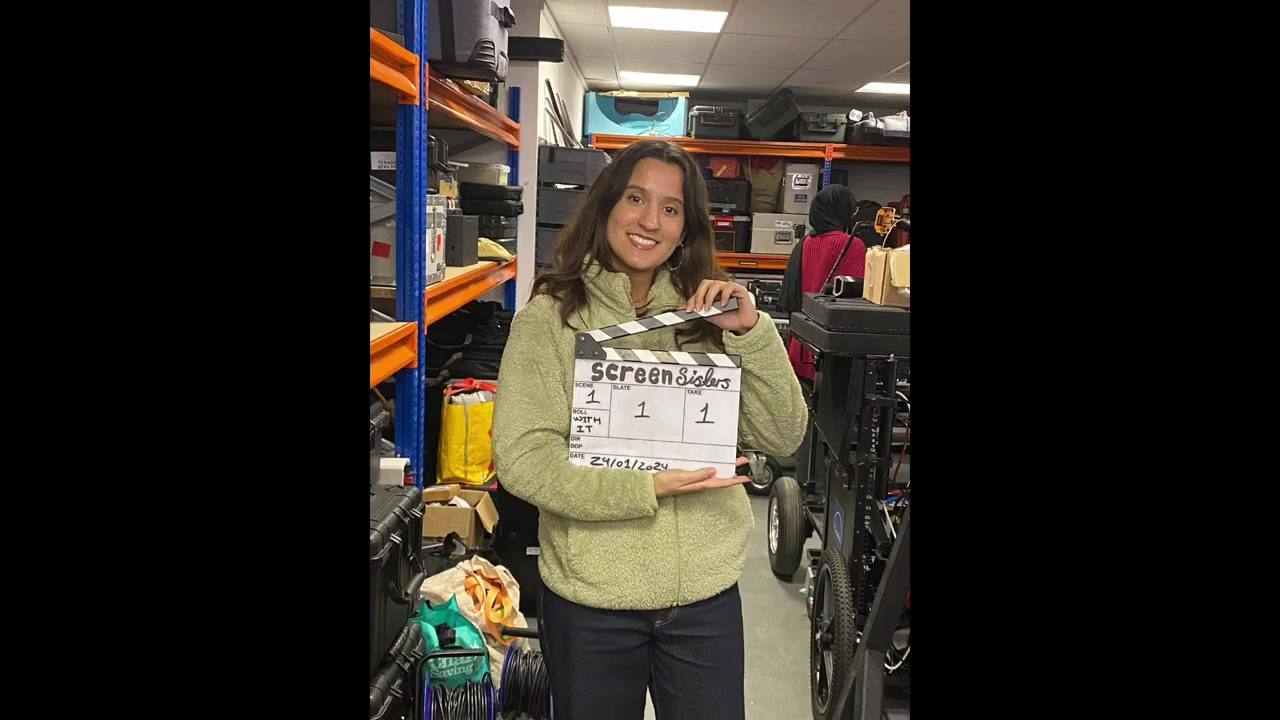 A woman holds up a clapperboard marked Screen Sisters, scene one, take one, standing in a kit store between racks of equipment and a dolly.