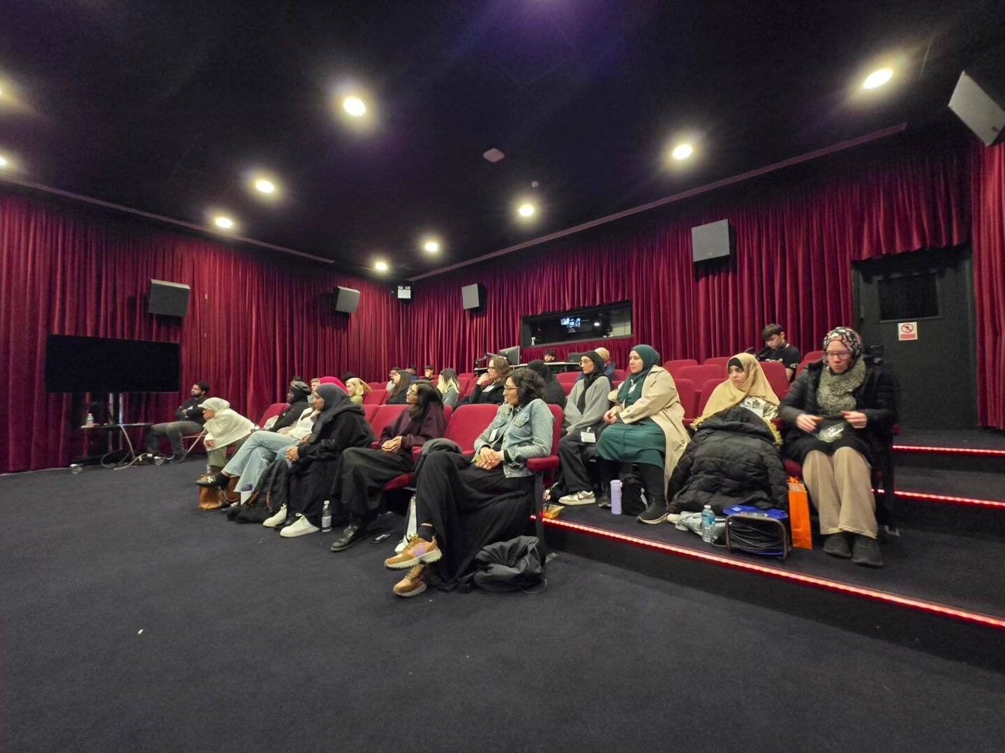 A small cinema auditorium with red curtains and tiered red seats, three rows of women sitting together with coats and bags around them, waiting for the lights to go down.