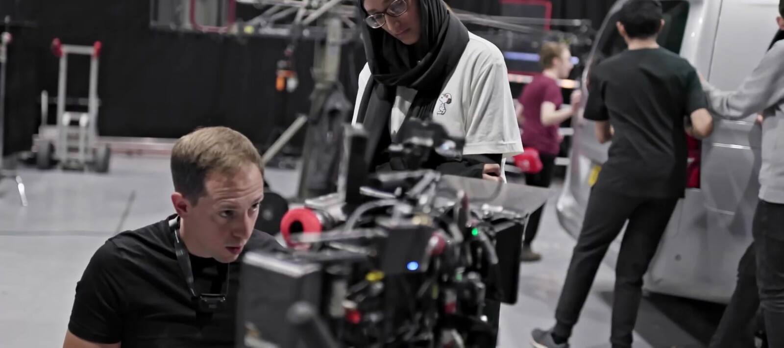 A woman in a hijab leans in over a built camera on a stand in a large studio space, a practitioner kneeling at the eyepiece side, a unit van and crew working behind them.