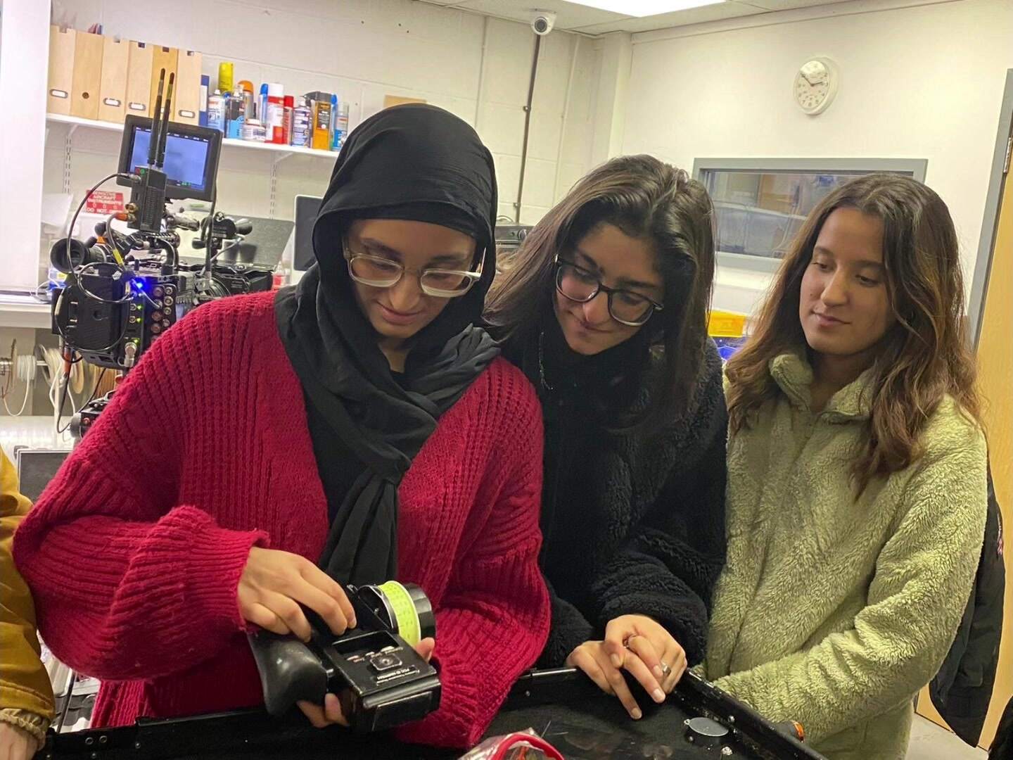 Three women stand together over a lens control handset on a camera trolley — one in a hijab with her hand on the wheel, two with uncovered hair watching what she is doing.