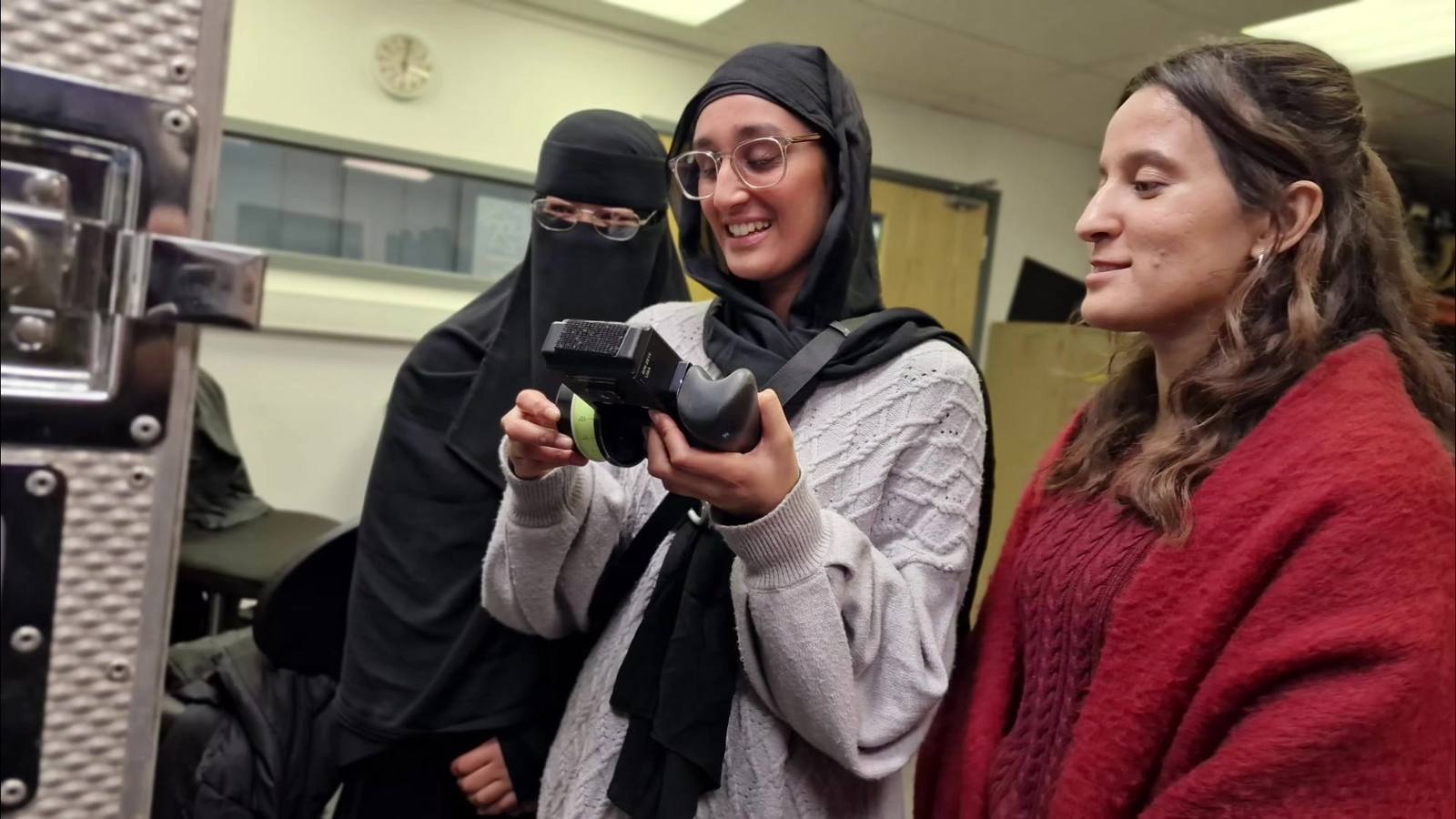 Three women laugh over a lens control handset in a kit room: one in a niqab, one in a hijab holding the handset, one with uncovered hair beside her.