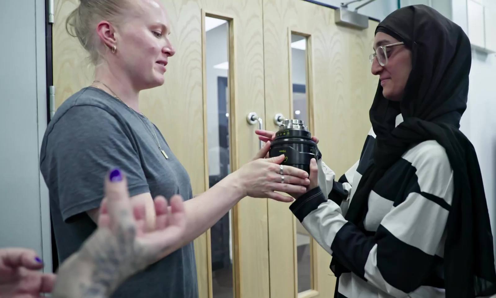 A woman with blonde hair and tattooed forearms passes a prime lens into the hands of a woman in a hijab and glasses, both looking at the lens as it changes hands.