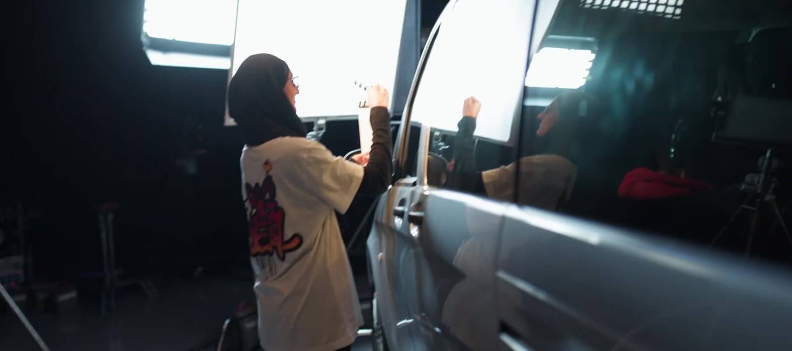 A woman in a hijab stands between two large LED panels set against the window of a car on a darkened stage, adjusting the light falling on the glass.