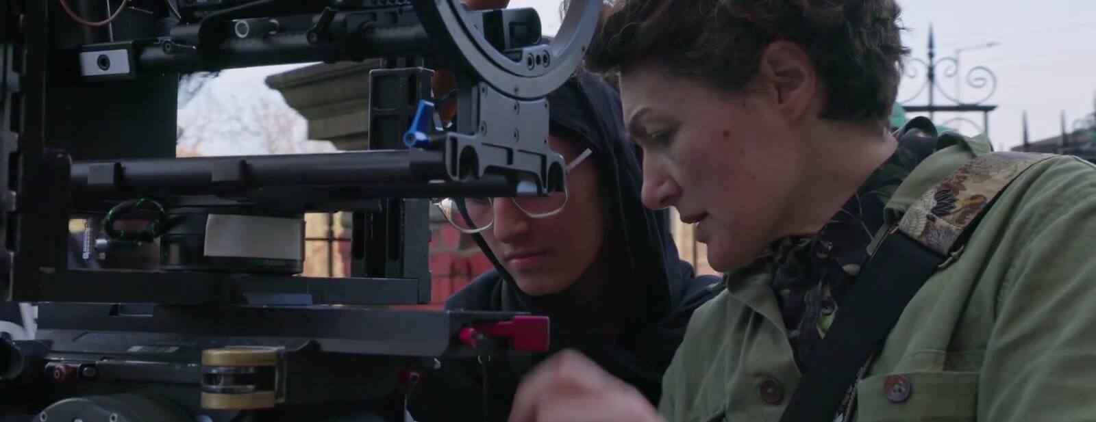 A woman with short uncovered hair and a woman in a hijab work side by side on a cinema camera rigged on a stabilising arm, both looking into the body as one of them reaches in to the lens.