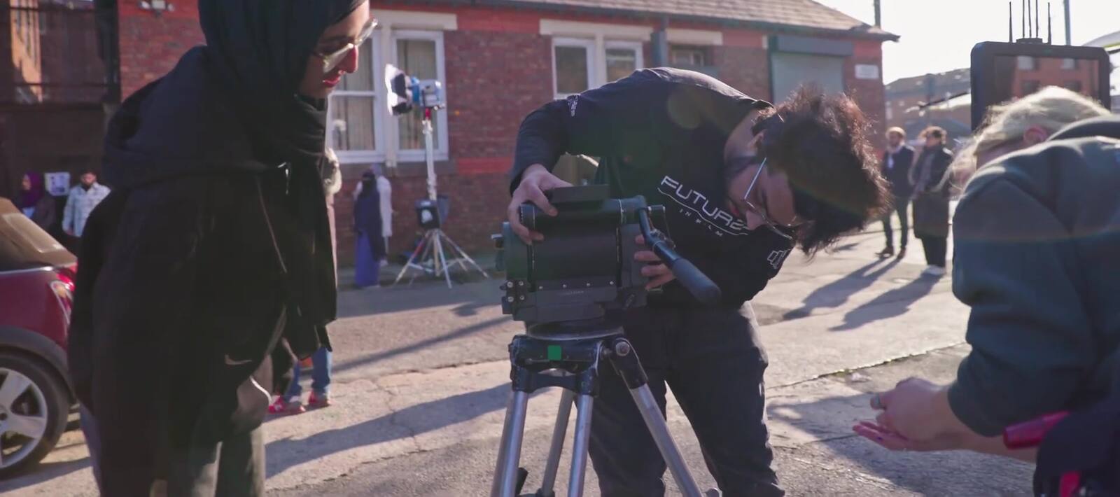 A camera unit at work in the middle of a street of red-brick terraced houses: a woman in a hijab at frame left, a crew member bent over the camera on its legs, and a woman with blonde hair crouching beside the tripod.