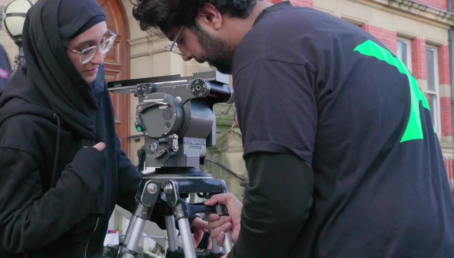 A woman in a hijab and a crew member lean over a fluid head on a tripod on a pavement, setting the legs outside a red-brick civic building.