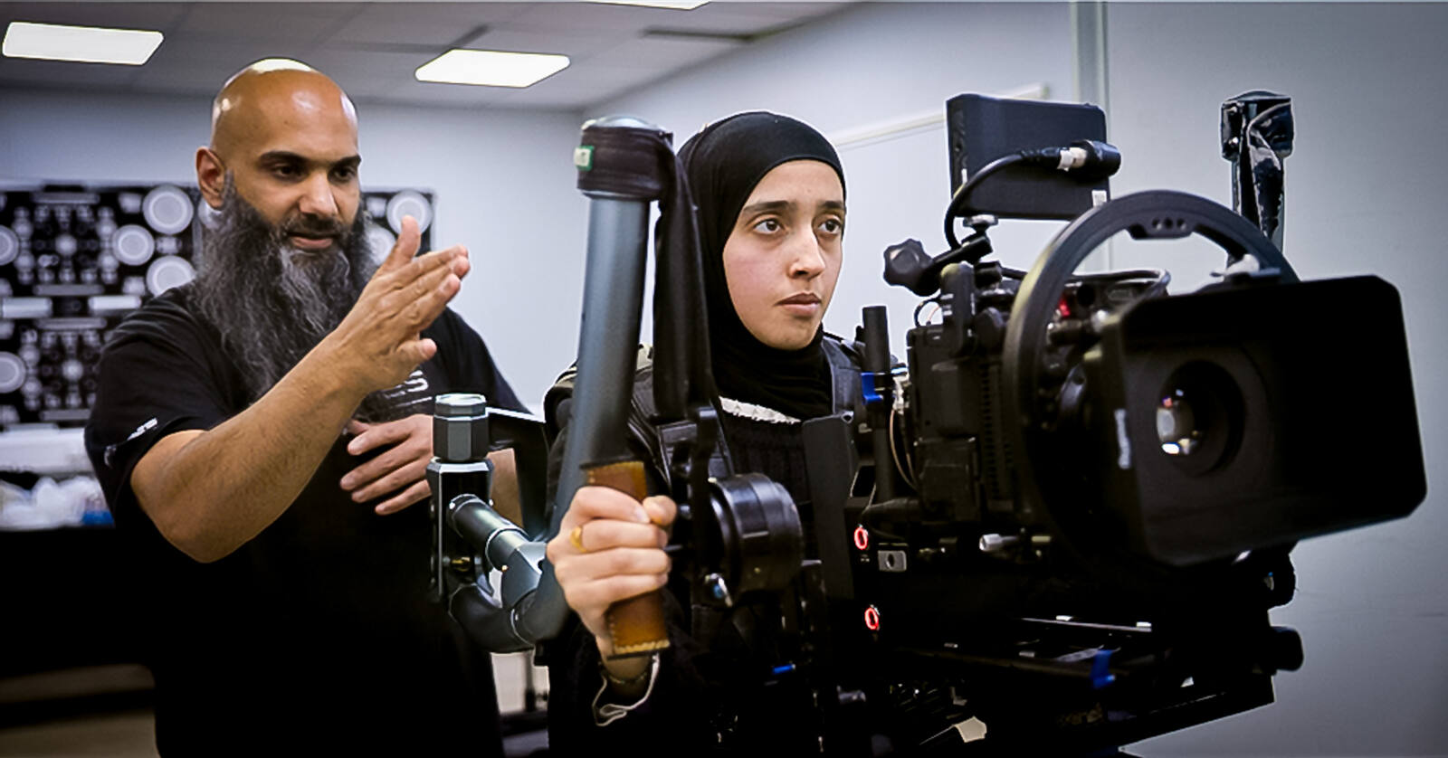 A woman in a hijab holds a built cinema camera on a body-mounted support rig, both hands on the arm, while a practitioner beside her talks through the movement with his hand raised.