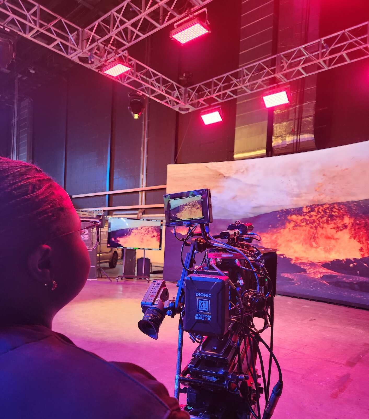 Looking over an operator's shoulder on a sound stage: a camera on a remote head faces a curved LED volume showing a lava landscape, with LED fixtures on a lighting truss overhead lighting the floor red.