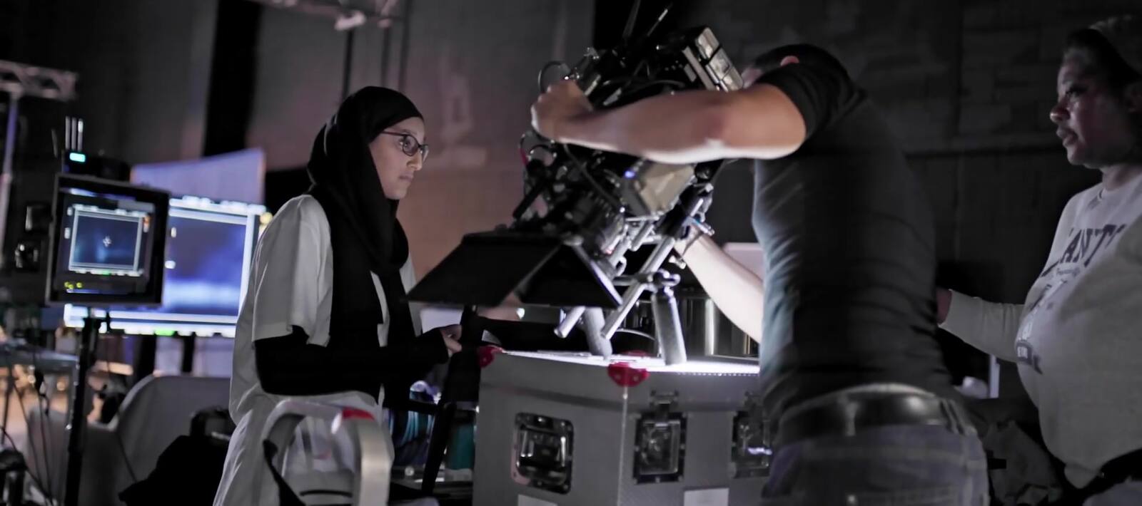 On a dark stage, a woman in a hijab watches a monitor at the side of a camera mounted on a remote head while a practitioner reaches across the rig to set it.