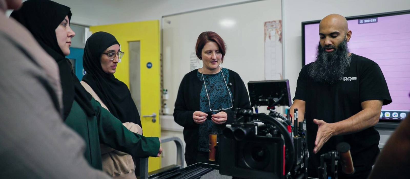 A practitioner talks a group through a built camera in a school classroom: two women in hijabs at frame left, a teacher with short red hair holding her glasses in the middle, a whiteboard and a display screen behind.