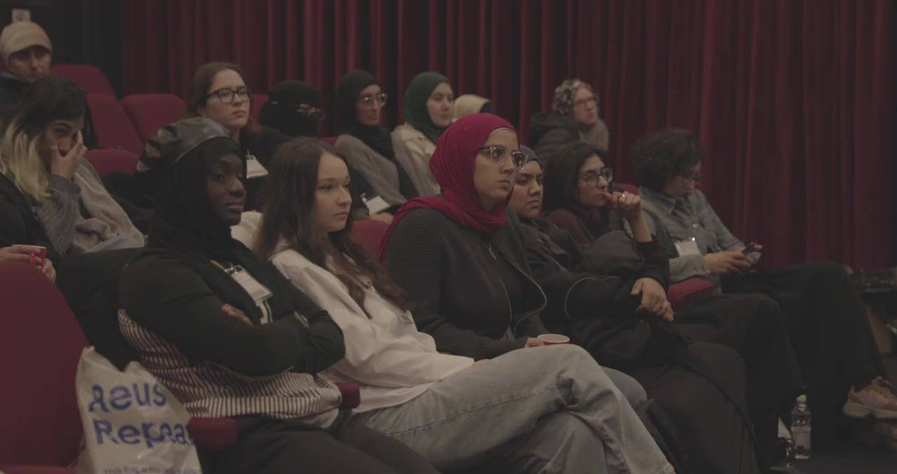 An audience watches a screening from red seats, faces lit by the screen: women in hijabs and niqabs, women with uncovered hair, sitting together across four rows.