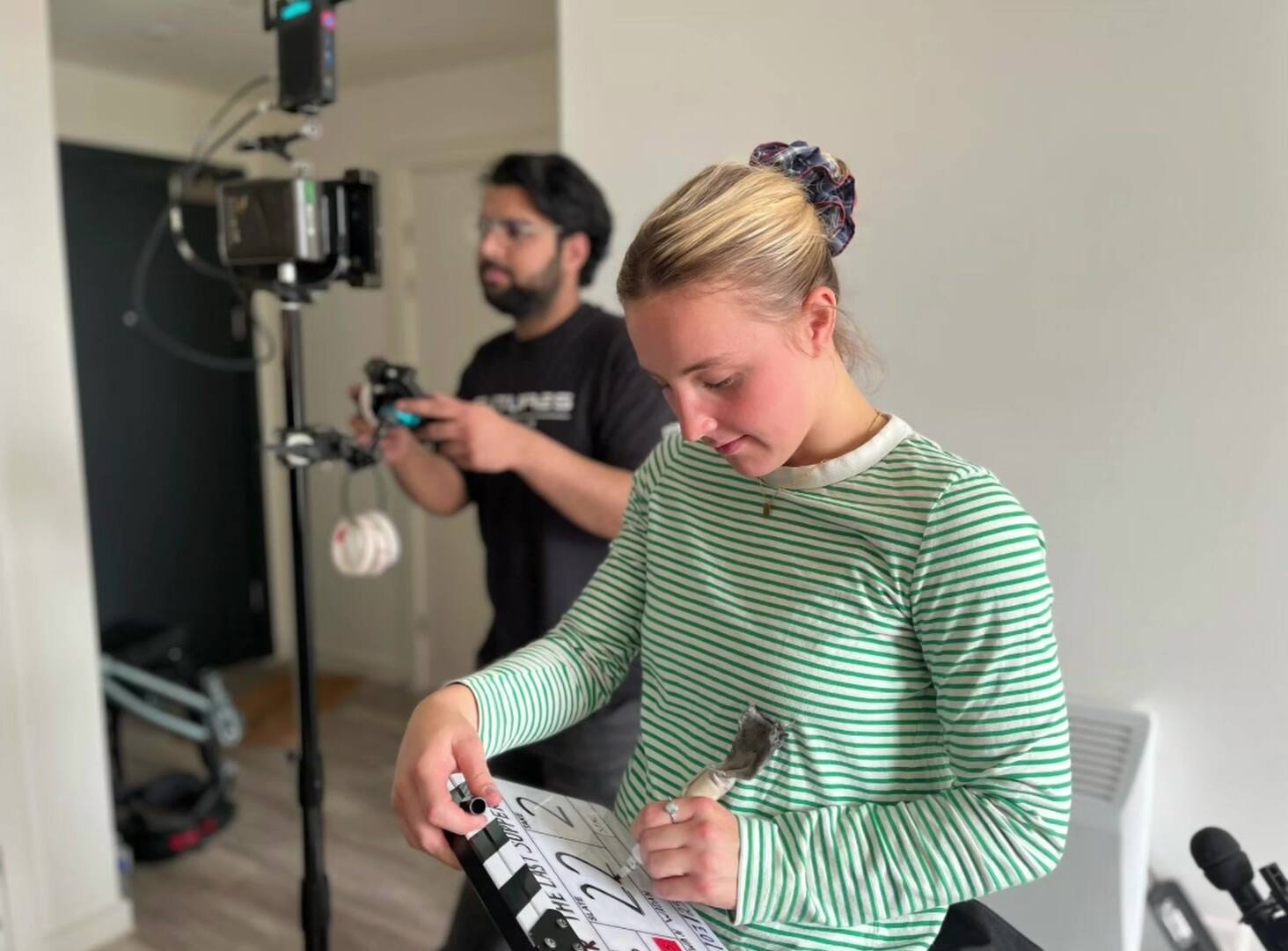 A woman with blonde hair tied up fills in a clapperboard with a marker while a crew member behind her sets a small camera on a stand in a bright room.