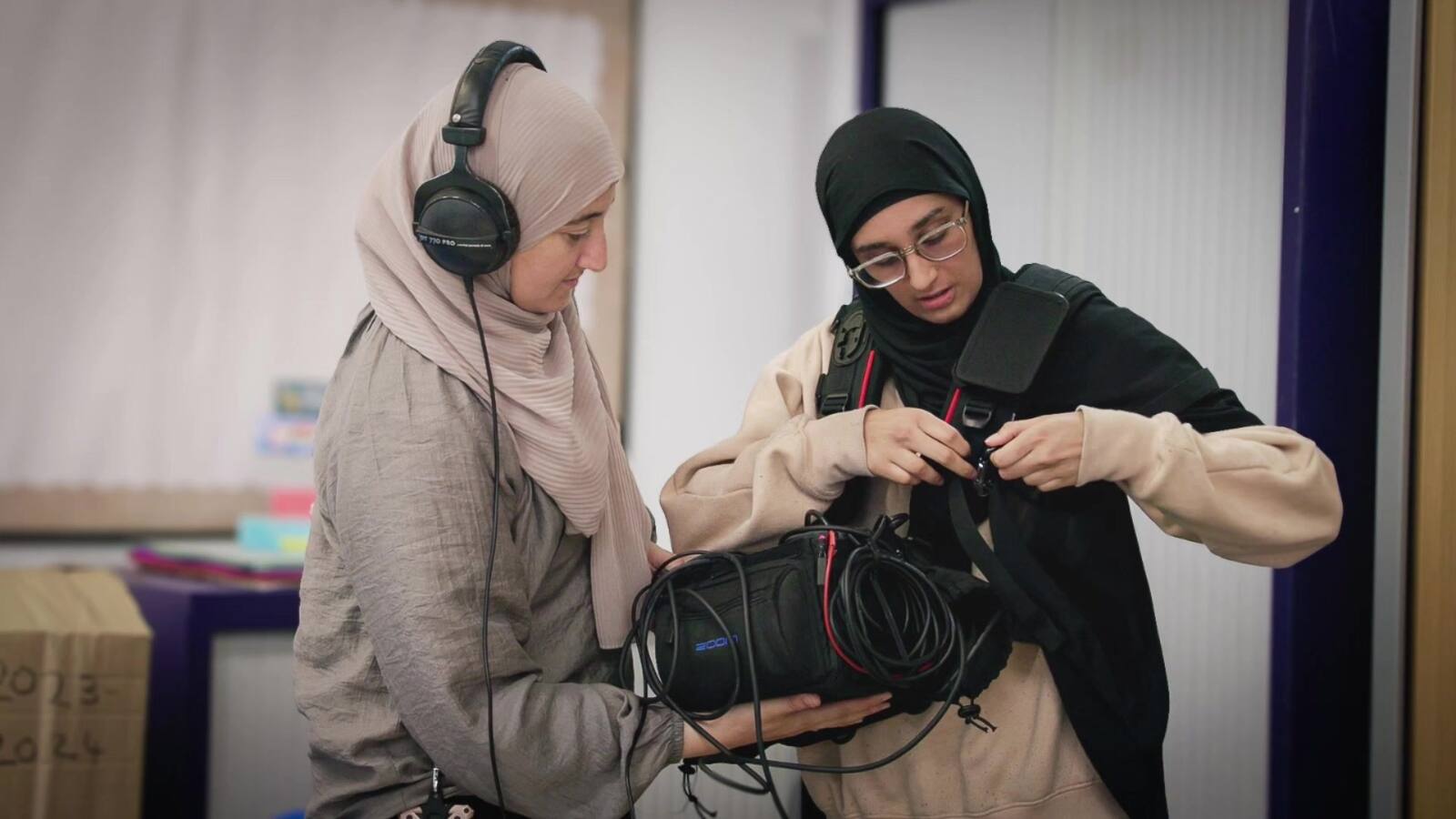 Two women rig a sound bag between them: one in a hijab wearing headphones holds the recorder steady, the other threads the harness over her shoulder.