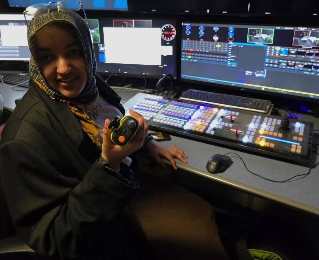 A woman in a patterned headscarf sits at a vision mixing desk in a gallery, one hand on the panel and a lens control handset in the other, the multiviewer and preview monitors lit in front of her.